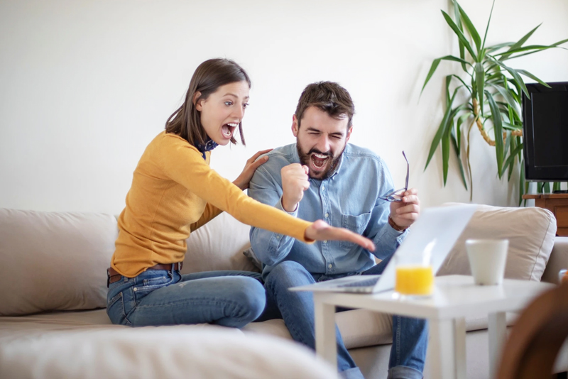 Excited couple looking at laptop while sitting on living room couch